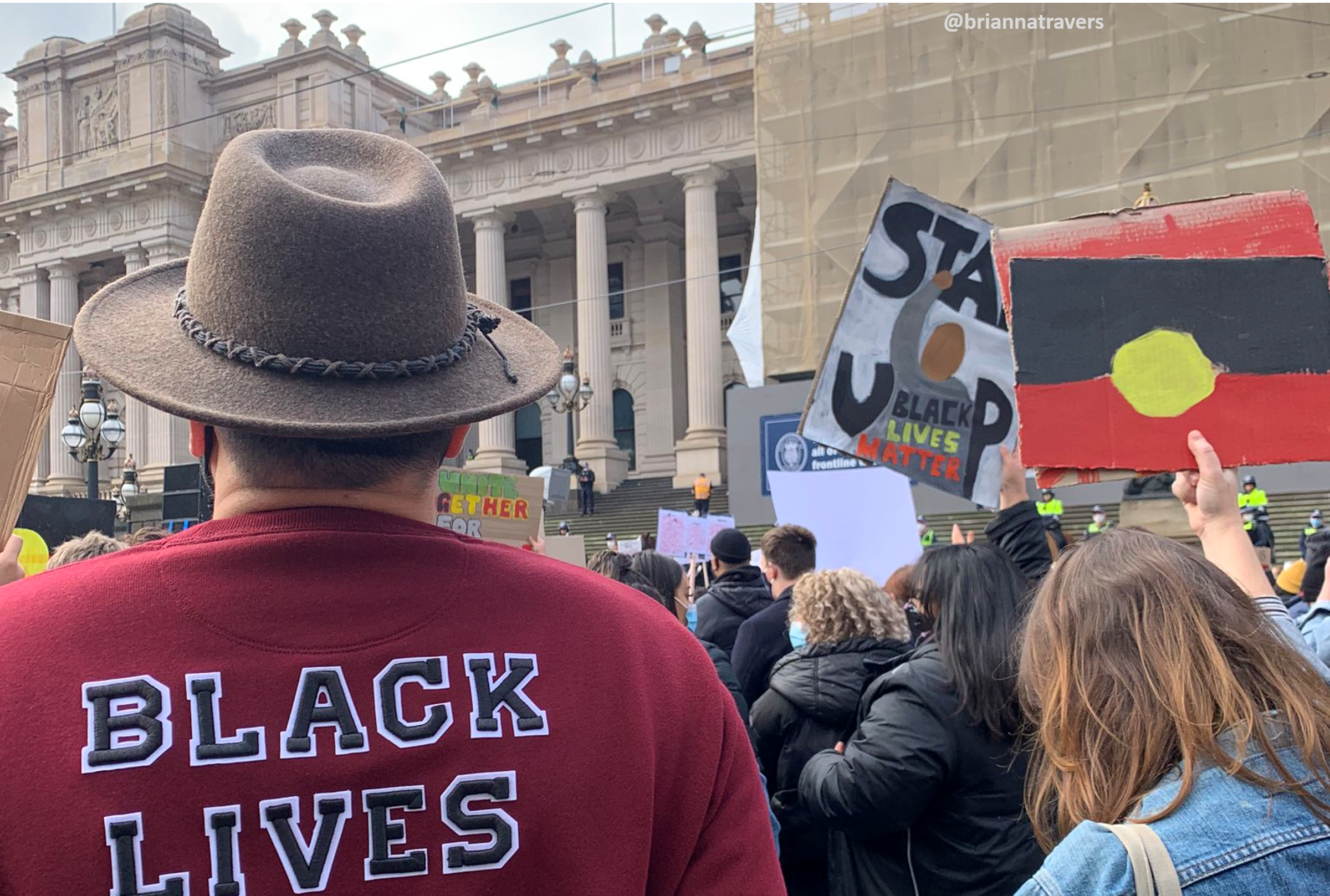 The backs of people's heads at a Black Lives Matter outside the Victorian Parliament. A woman holds a placard with the Aboriginal flag, a man in an akubra has 'BLACK LIVES MATTER' across the back of his jumper. 